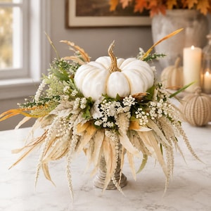 May include: A white decorative pumpkin with a gold stem sits atop a pedestal base, surrounded by an arrangement of cream, beige, and green faux foliage, including feathery grasses, small white berries, and various textured autumn leaves.