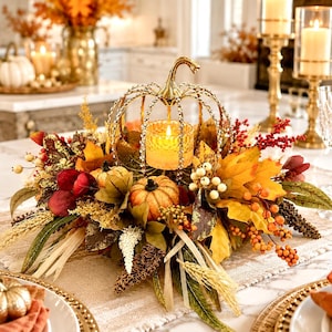 May include: A decorative centerpiece featuring a gold-toned metal pumpkin frame with crystal accents, surrounding a lit votive candle. The arrangement includes artificial autumn leaves in shades of orange, yellow, and red, small pumpkins, and berries.