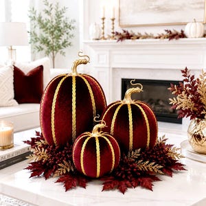 May include: Three velvet pumpkins in a deep burgundy color, each featuring vertical gold braided trim and a metallic gold stem. The pumpkins are arranged in a cluster, resting on a base of dark red faux leaves and gold-toned foliage accents.