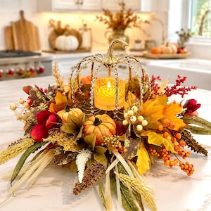 May include: A decorative centerpiece featuring a gold-toned metal pumpkin frame with crystal accents, holding a glowing votive candle. It is surrounded by an arrangement of artificial autumn foliage, including orange pumpkins, red berries, and wheat.