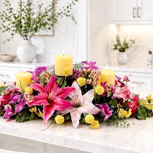 May include: A floral centerpiece with three lit yellow candles surrounded by pink lilies, purple orchids, and yellow flowers. The arrangement sits on a white countertop in a kitchen setting.