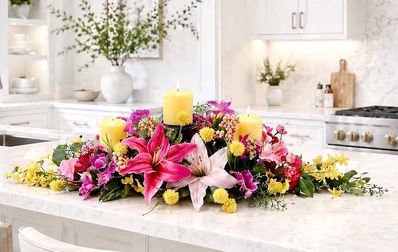 May include: A floral centerpiece with three lit yellow candles surrounded by pink lilies, purple orchids, and yellow flowers. The arrangement sits on a white countertop in a kitchen setting.
