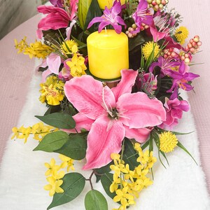 May include: A floral centerpiece featuring vibrant pink lilies, purple orchids, and yellow flowers. Two yellow pillar candles are nestled among the blooms. Green leaves and berries add to the arrangement's natural aesthetic.