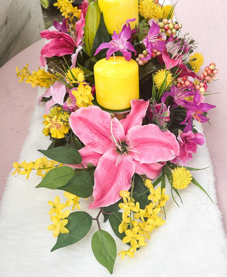 May include: A floral centerpiece featuring vibrant pink lilies, purple orchids, and yellow flowers. Two yellow pillar candles are nestled among the blooms. Green leaves and berries add to the arrangement's natural aesthetic.