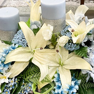 Puede incluir: Un centro de mesa con tres velas azules con detalles de purpurina, rodeadas de lirios blancos y flores azules.