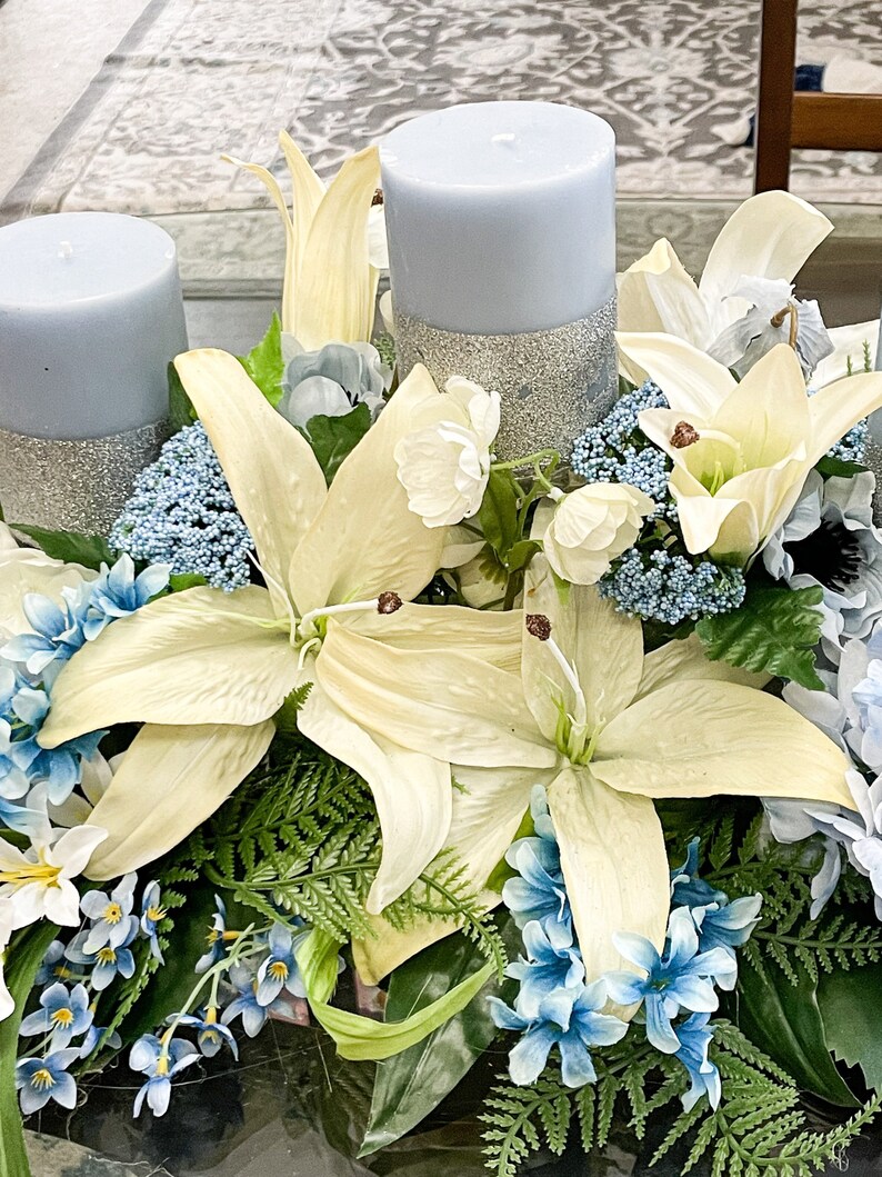 Puede incluir: Un centro de mesa con tres velas azules con detalles de purpurina, rodeadas de lirios blancos y flores azules.