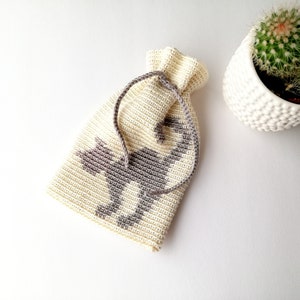 May include: A cream-colored, crocheted drawstring bag with a gray geometric design of a horse. The bag is lying on a white surface next to a white textured pot with a cactus.
