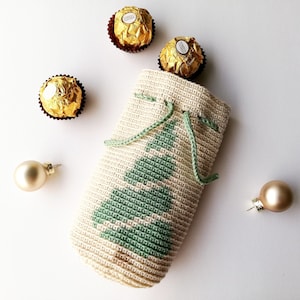 May include: A beige crocheted gift bag with a green Christmas tree design, drawstring closure, and green cord. The bag is surrounded by three gold-wrapped chocolate candies and two gold Christmas ornaments.