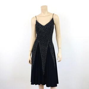 May include: A black knee-length dress on a mannequin. The dress features a V-neckline and spaghetti straps, both adorned with small, white embellishments. White embellishments also form a geometric pattern on the bodice and skirt.