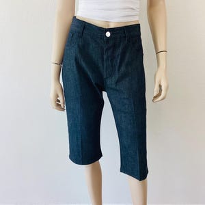 May include: Dark blue denim capri pants with a button closure and pockets. The capri pants are displayed on a mannequin. The pants are knee-length and have a classic, tailored design.