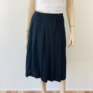 Op de afbeelding: Zwarte midi-rok met oogjes en een knoopsluiting. De rok wordt op een mannequin getoond. De rok heeft een hoge taille en een geplooid ontwerp. De rok is knielang en heeft een geschulpte zoom.