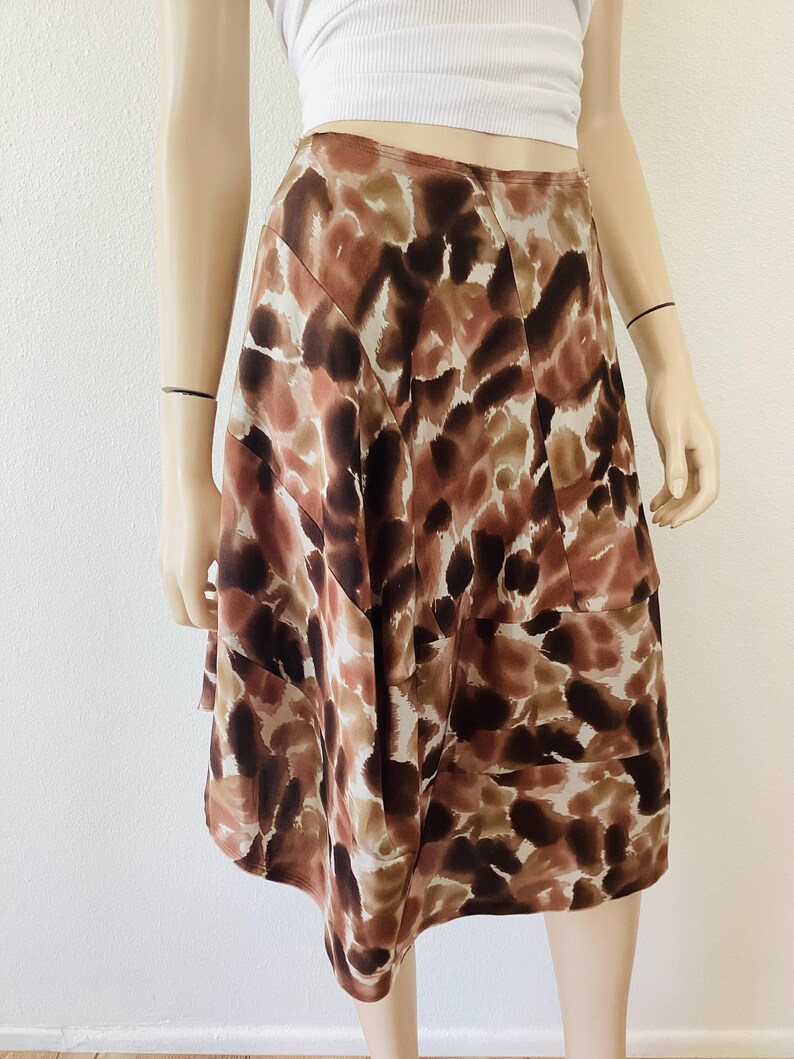 K&ouml;nnte beinhalten: Ein knielanger Rock mit einem abstrakten Muster in Braun, Beige und Wei&szlig;. Der Rock hat eine hohe Taille und eine ausgestellte Silhouette. Der Stoff scheint leicht und flie&szlig;end zu sein. Der Rock wird auf einer Schaufensterpuppe pr&auml;sentiert.