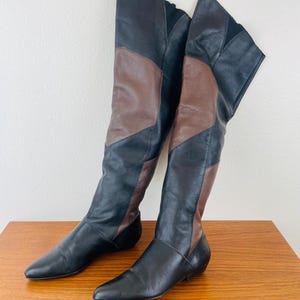 May include: A pair of knee-high boots in black and brown leather. The boots have a pointed toe and a low heel. The design features a color-blocked pattern with black and brown leather panels. The boots are displayed on a wooden surface.