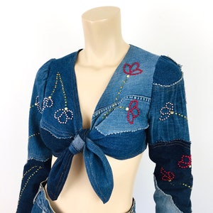 May include: A blue denim top with long sleeves and a tie front. The top is made of different pieces of denim sewn together and decorated with colorful rhinestones.