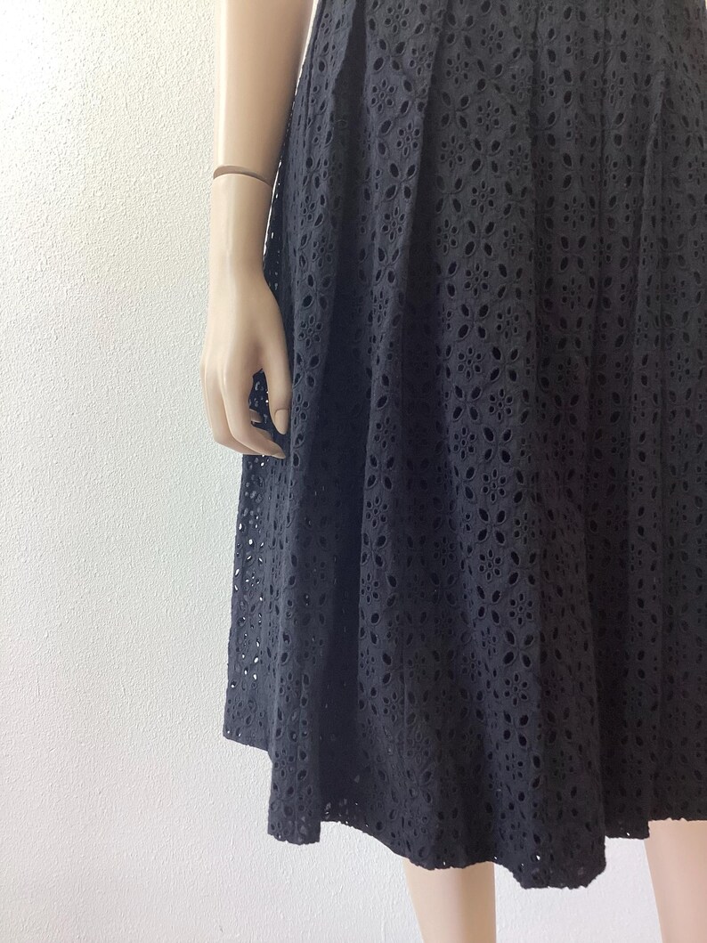 Op de afbeelding: Zwarte eyelet rok met een bloemenpatroon. De rok is geplooid en valt tot halverwege de kuit. De stof heeft een delicaat, openwerk ontwerp, waardoor een textuur ontstaat. De rok wordt op een mannequin getoond.