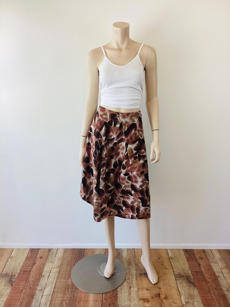 K&ouml;nnte beinhalten: Ein wei&szlig;es Tanktop kombiniert mit einem knielangen Rock. Der Rock hat ein abstraktes Muster in Braun, Wei&szlig; und Beige. Die Kleidung wird auf einer Schaufensterpuppe vor einer wei&szlig;en Wand und einem Holzboden pr&auml;sentiert.