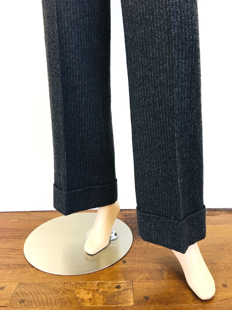 Puede incluir: Un par de pantalones de rayas finas de color gris oscuro con dobladillos. Los pantalones est&aacute;n en un maniqu&iacute; y se ve la parte inferior de los pantalones.