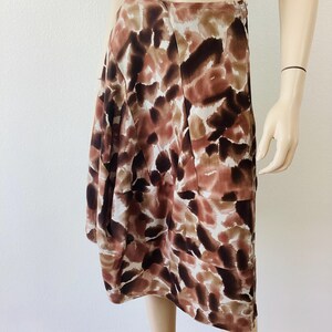 K&ouml;nnte beinhalten: Knie-langer Rock mit einem abstrakten Muster in Braun, Beige und Wei&szlig;. Der Rock ist aus leichtem Stoff gefertigt und hat ein plissiertes Design. Der Rock wird auf einer Schaufensterpuppe pr&auml;sentiert.