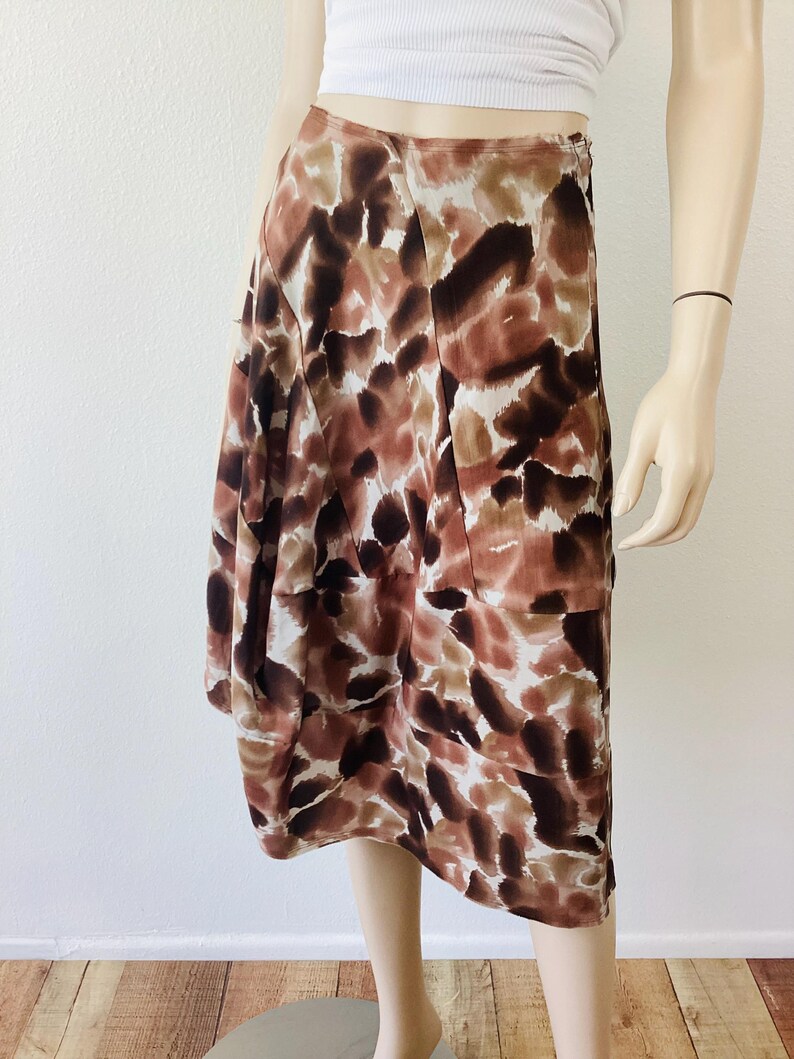 K&ouml;nnte beinhalten: Knie-langer Rock mit einem abstrakten Muster in Braun, Beige und Wei&szlig;. Der Rock ist aus leichtem Stoff gefertigt und hat ein plissiertes Design. Der Rock wird auf einer Schaufensterpuppe pr&auml;sentiert.