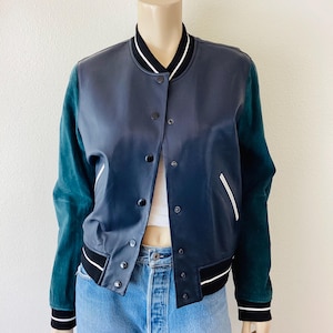 May include: A two-tone varsity jacket with a navy blue body and teal green sleeves. The jacket features black and white striped trim on the collar, cuffs, and hem. It has a button-up front and a classic bomber jacket style.
