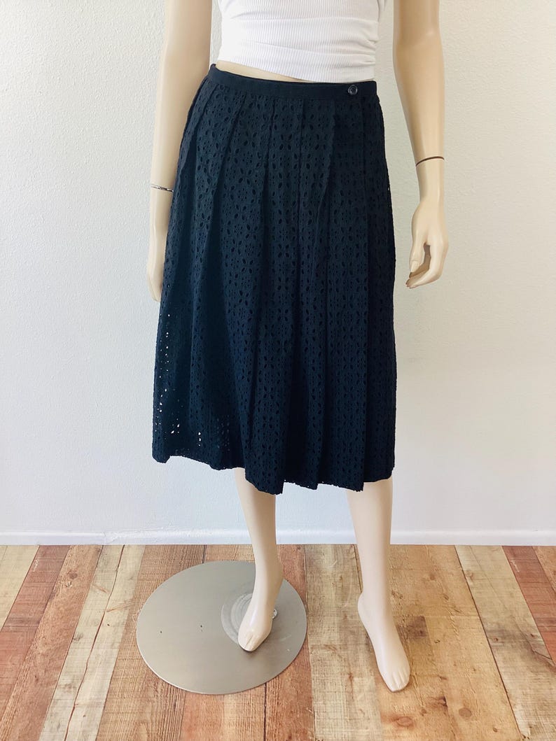 Op de afbeelding: Zwarte midi-rok met oogjes en een knoopsluiting. De rok wordt op een mannequin getoond. De rok heeft een hoge taille en een geplooid ontwerp. De rok is knielang en heeft een geschulpte zoom.