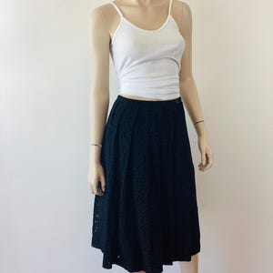 Op de afbeelding: Een mannequin draagt een witte geribbelde tanktop en een zwarte midi-rok met oogjes. De rok heeft een knoopsluiting en een uitlopende vorm. De outfit is eenvoudig en klassiek, geschikt voor diverse gelegenheden.