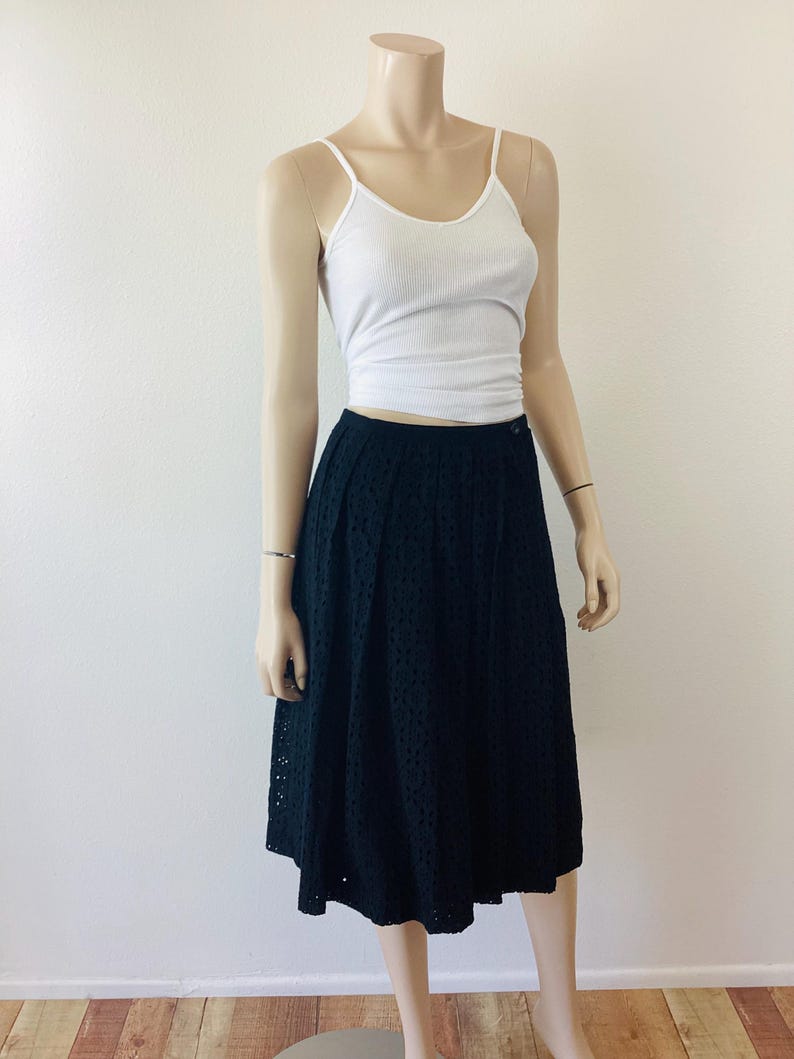 Op de afbeelding: Een mannequin draagt een witte geribbelde tanktop en een zwarte midi-rok met oogjes. De rok heeft een knoopsluiting en een uitlopende vorm. De outfit is eenvoudig en klassiek, geschikt voor diverse gelegenheden.