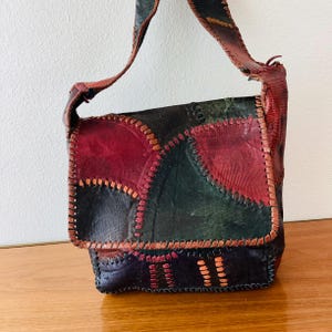 May include: A vintage patchwork leather shoulder bag. The bag features a flap closure and a long strap. The leather is a mix of colors, including red, green, and black, with visible stitching. The bag is likely handmade.