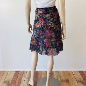 May include: A-line skirt with a floral pattern in shades of purple, pink, and yellow. The skirt has a ruffled hem and is made of a sheer fabric. The mannequin is also wearing a white vest top.