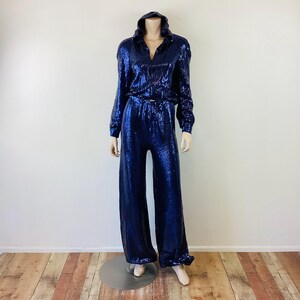 May include: A navy blue sequined jumpsuit with a hooded top and wide-leg pants. The top has a V-neck and long sleeves. The pants are high-waisted and include a matching belt. The outfit is displayed on a mannequin.