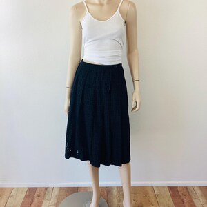 Op de afbeelding: Een zwarte midi-rok met oogjes gecombineerd met een witte geribbelde tanktop op een mannequin. De rok heeft een hoge taille en een uitlopend silhouet. De tanktop heeft dunne bandjes. De mannequin staat op een metalen standaard, tegen een witte muur en een houten vloer.