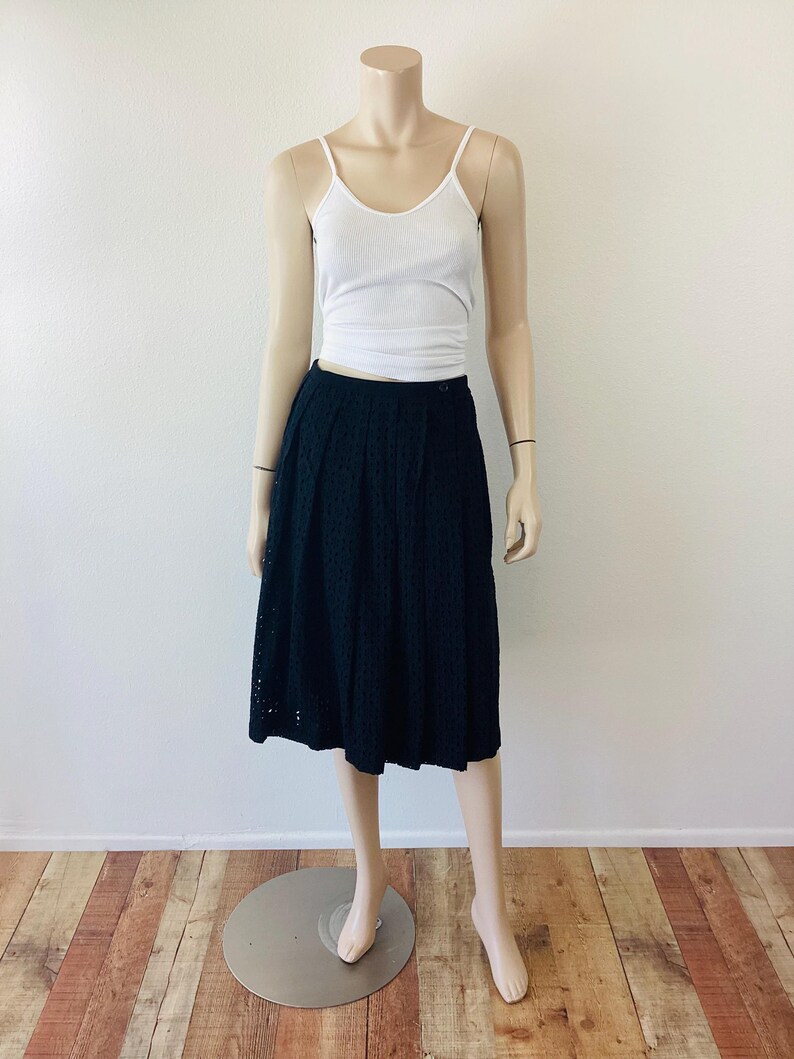 Op de afbeelding: Een zwarte midi-rok met oogjes gecombineerd met een witte geribbelde tanktop op een mannequin. De rok heeft een hoge taille en een uitlopend silhouet. De tanktop heeft dunne bandjes. De mannequin staat op een metalen standaard, tegen een witte muur en een houten vloer.