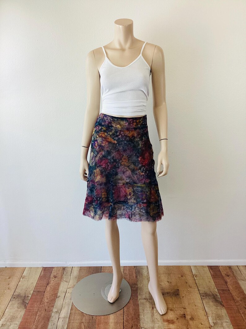 May include: A mannequin wearing a white vest top and a knee-length skirt. The skirt has a colourful floral pattern with shades of blue, purple, and pink. The skirt has a ruffled hemline.
