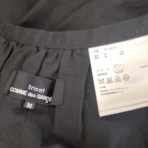 Op de afbeelding: Close-up van een zwart kledingstuk met een label van tricot COMME des GAR&Ccedil;ONS. Het label is zwart met witte tekst en de maat is M. Een wit label met Japanse tekst en verzorgingssymbolen is ook zichtbaar.