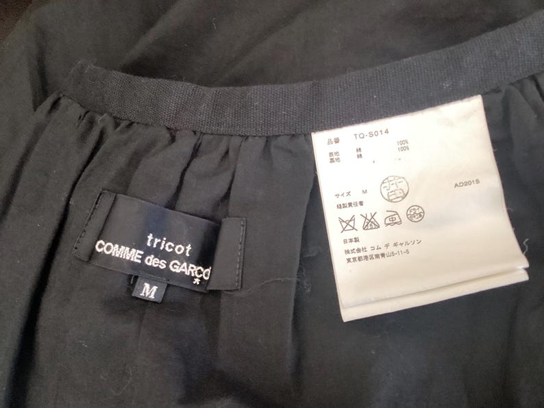 Op de afbeelding: Close-up van een zwart kledingstuk met een label van tricot COMME des GAR&Ccedil;ONS. Het label is zwart met witte tekst en de maat is M. Een wit label met Japanse tekst en verzorgingssymbolen is ook zichtbaar.