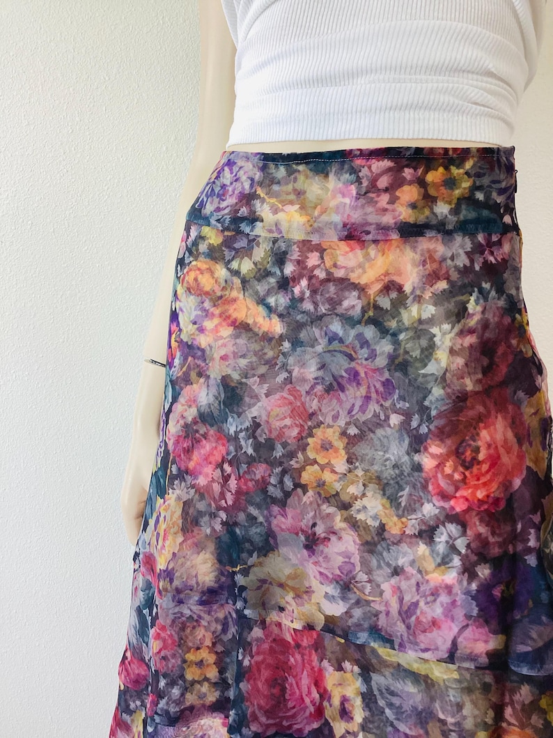 May include: A colourful floral print skirt in shades of pink, purple, yellow, and orange. The skirt has a flowing, layered design and is made of a sheer fabric. The skirt is part of a clothing collection.