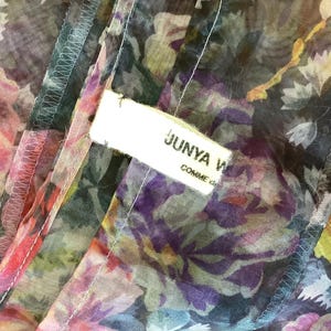 May include: Close-up of a floral patterned fabric with a white label that reads "JUNYA WATANABE COMME des GAR&Ccedil;ONS". The fabric features a mix of pink, purple, yellow, and green floral designs on a dark background. The edges are stitched with white thread.