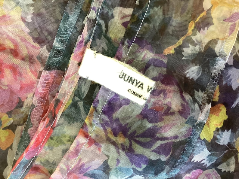 May include: Close-up of a floral patterned fabric with a white label that reads "JUNYA WATANABE COMME des GAR&Ccedil;ONS". The fabric features a mix of pink, purple, yellow, and green floral designs on a dark background. The edges are stitched with white thread.