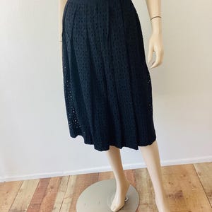 Op de afbeelding: Zwarte midi-rok met oogjes. De rok heeft een geplooid ontwerp en een zoom tot op de knie. De rok wordt op een mannequin getoond. De rok is gemaakt van een lichte stof, perfect voor warm weer.