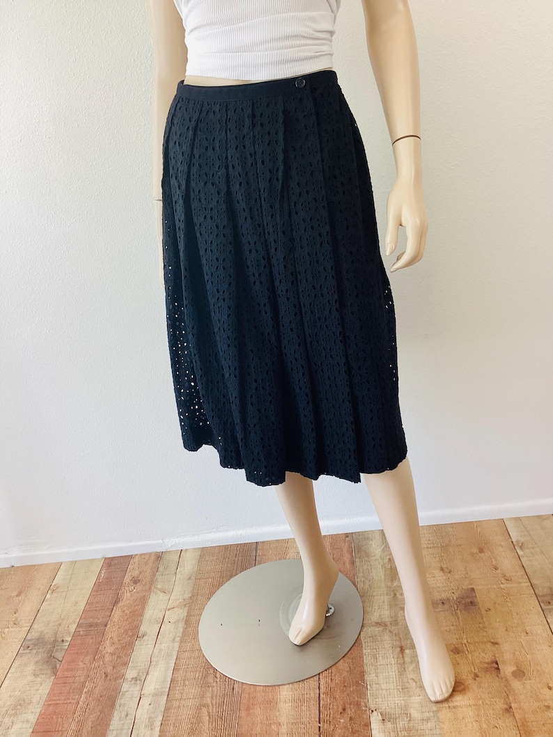 Op de afbeelding: Zwarte midi-rok met oogjes. De rok heeft een geplooid ontwerp en een zoom tot op de knie. De rok wordt op een mannequin getoond. De rok is gemaakt van een lichte stof, perfect voor warm weer.