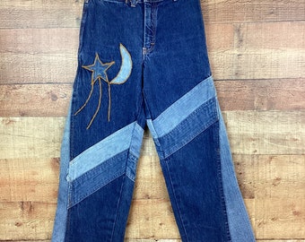 May include: Blue denim jeans with a crescent moon and star design on the left leg. The jeans have a patchwork design on the legs, with different shades of blue denim.
