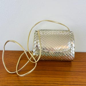 May include: A small, gold-colored handbag with a textured, scale-like pattern. The bag has a rectangular shape with a curved flap and a thin, gold shoulder strap. The bag is sitting on a wooden surface.