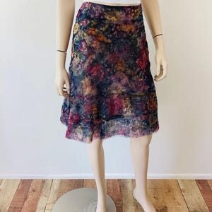 May include: A floral patterned skirt in shades of purple, pink, yellow, and blue. The skirt is knee-length and has a layered design. It is displayed on a mannequin. The skirt is made of a sheer fabric.