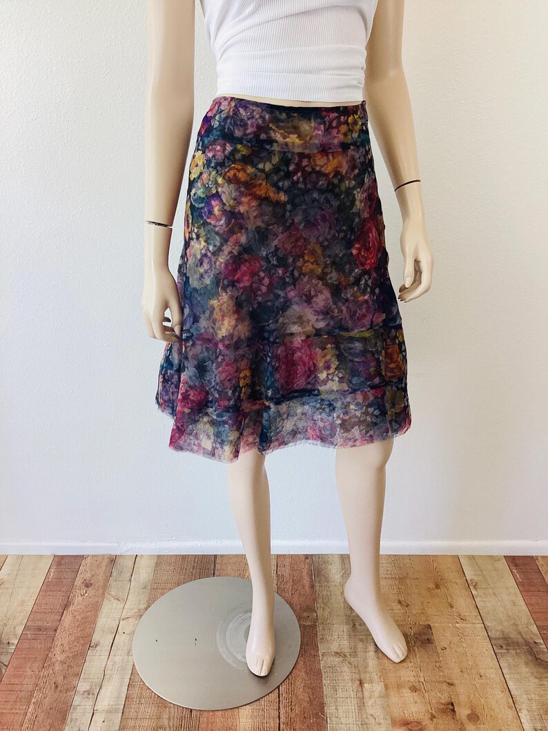 May include: A floral patterned skirt in shades of purple, pink, yellow, and blue. The skirt is knee-length and has a layered design. It is displayed on a mannequin. The skirt is made of a sheer fabric.