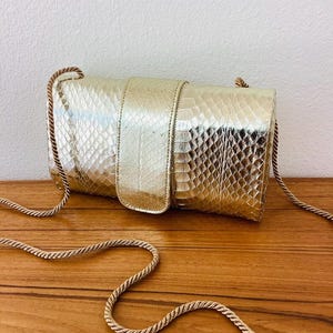 May include: A gold clutch with a snakeskin pattern and a matching strap. The bag has a flap closure and a gold strap. The bag is sitting on a wooden surface.