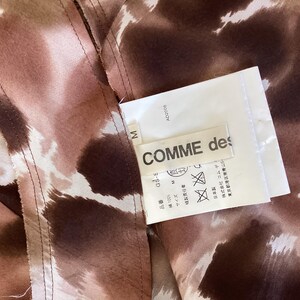 K&ouml;nnte beinhalten: Nahaufnahme eines Kleidungsst&uuml;cks mit braunem und wei&szlig;em Muster. Ein wei&szlig;es Etikett mit dem Text "COMME des" ist angebracht. Das Kleidungsst&uuml;ck hat sichtbare N&auml;hte.