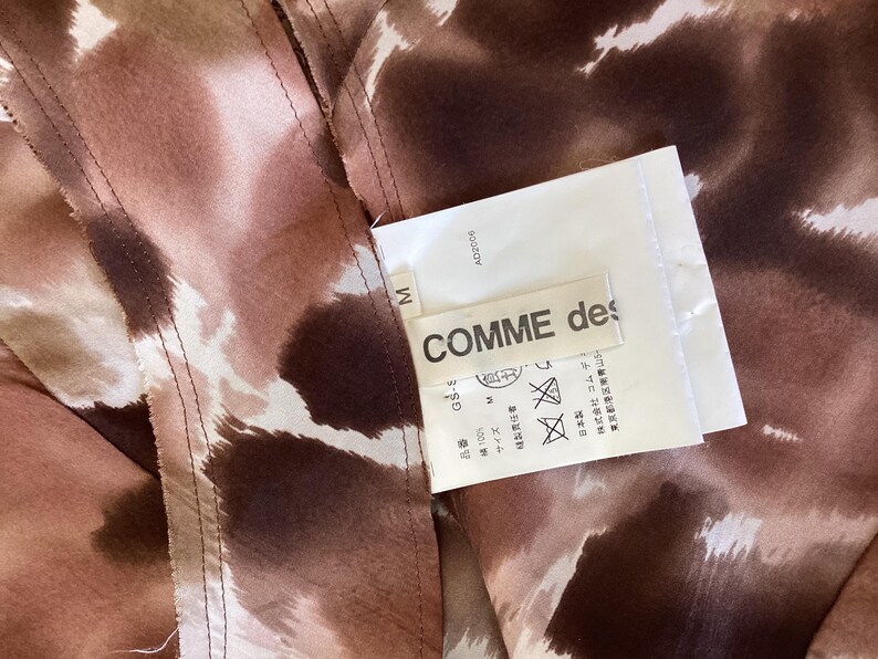K&ouml;nnte beinhalten: Nahaufnahme eines Kleidungsst&uuml;cks mit braunem und wei&szlig;em Muster. Ein wei&szlig;es Etikett mit dem Text "COMME des" ist angebracht. Das Kleidungsst&uuml;ck hat sichtbare N&auml;hte.