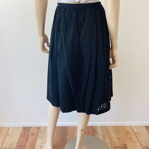 Op de afbeelding: Zwarte midi-rok met oogjes. De rok heeft een hoge taille en een uitlopend silhouet. De stof is effen zwart met een all-over oogjespatroon. De rok wordt op een mannequin getoond.