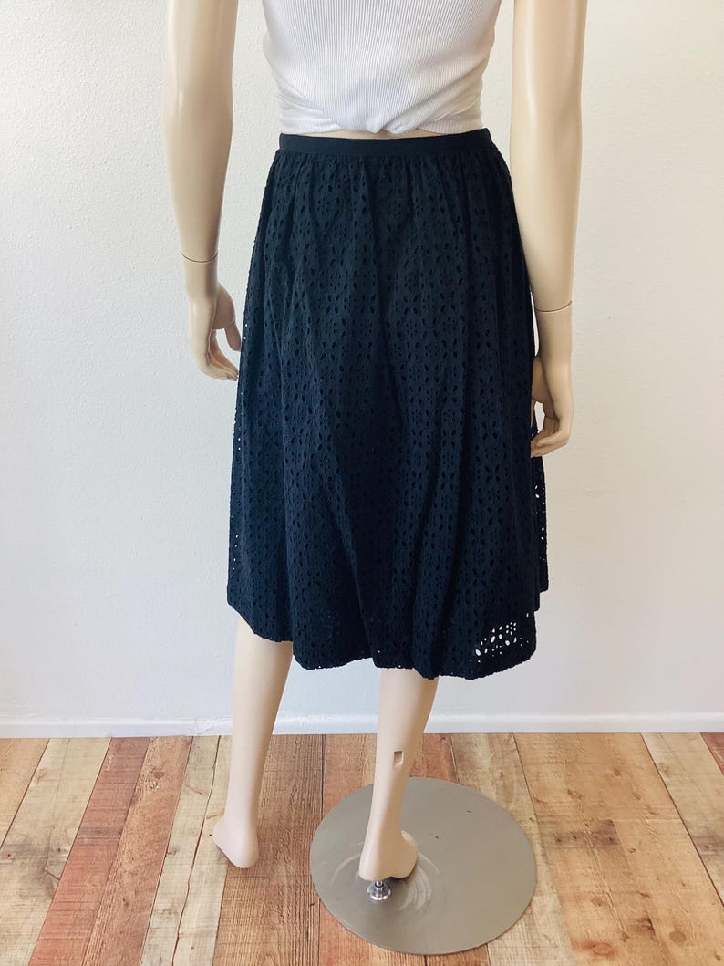 Op de afbeelding: Zwarte midi-rok met oogjes. De rok heeft een hoge taille en een uitlopend silhouet. De stof is effen zwart met een all-over oogjespatroon. De rok wordt op een mannequin getoond.