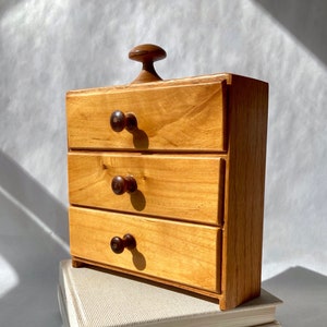 May include: A small, wooden chest of drawers with three drawers and a top knob. The chest is light brown with dark brown knobs and sits atop two stacked books with off-white covers.